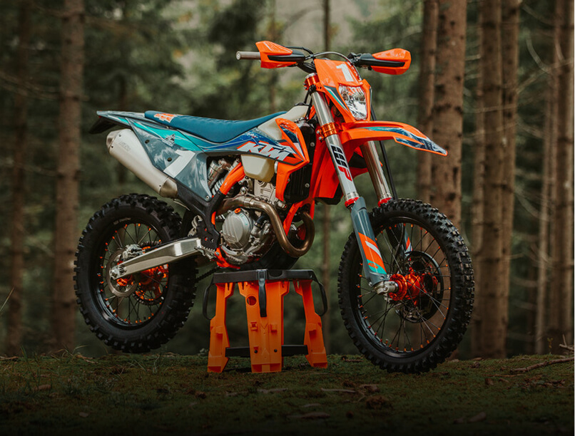 An orange dirt bike parked among trees in a wooded area, showcasing its vibrant color against the natural backdrop.